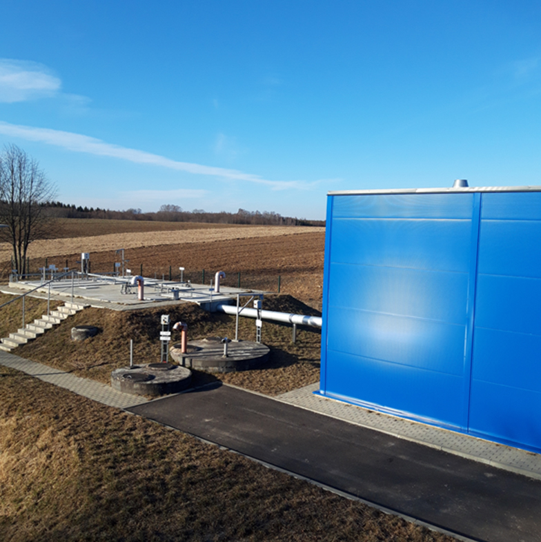 Nuotekų valymo įrenginiai: rezervuarai, vamzdynai ir mėlynas technologinis pastatas.
