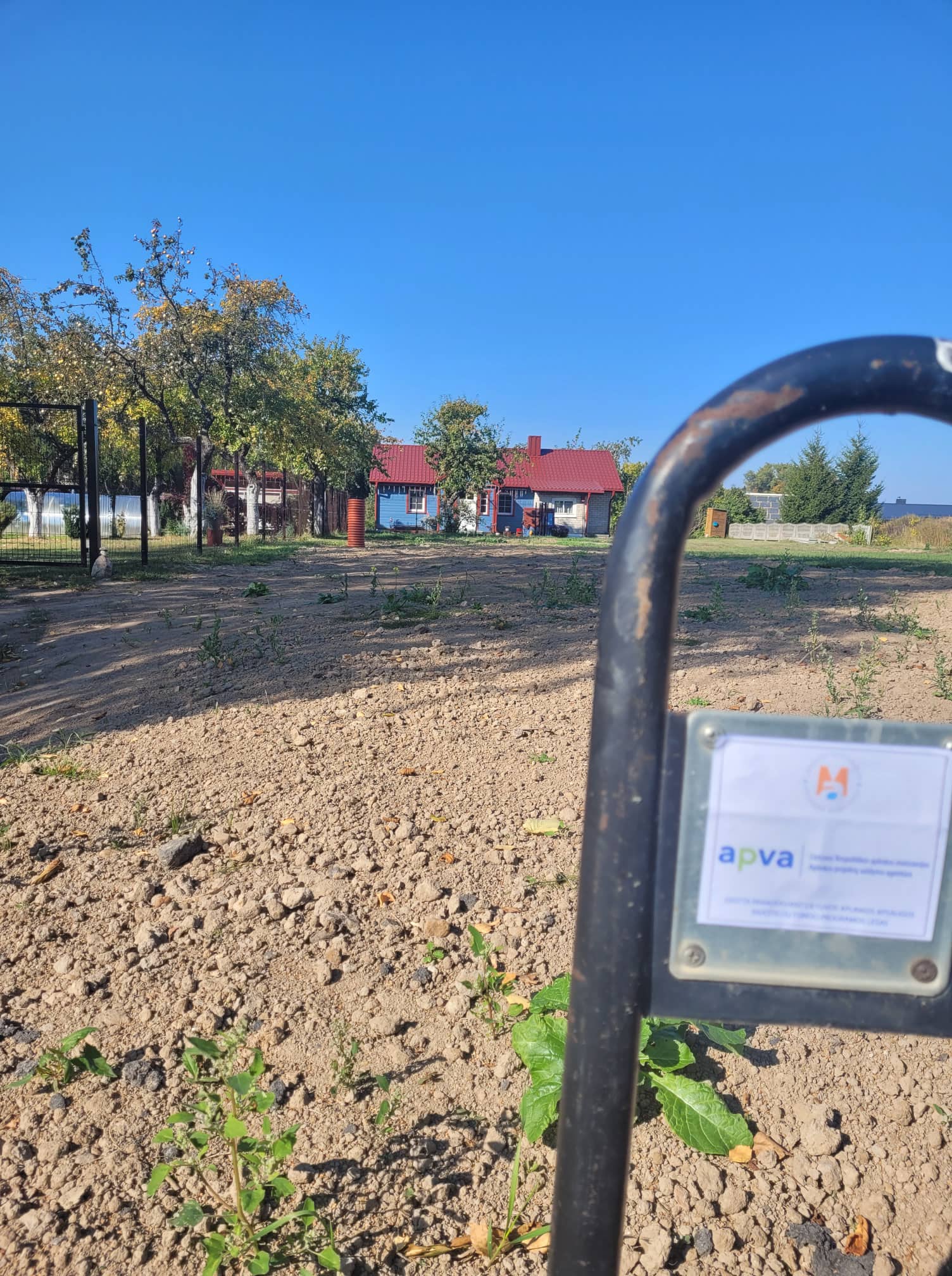 Projekto vietos nuotrauka: kiemas po žemės darbų ir APVA finansavimo ženklas ant vartų.