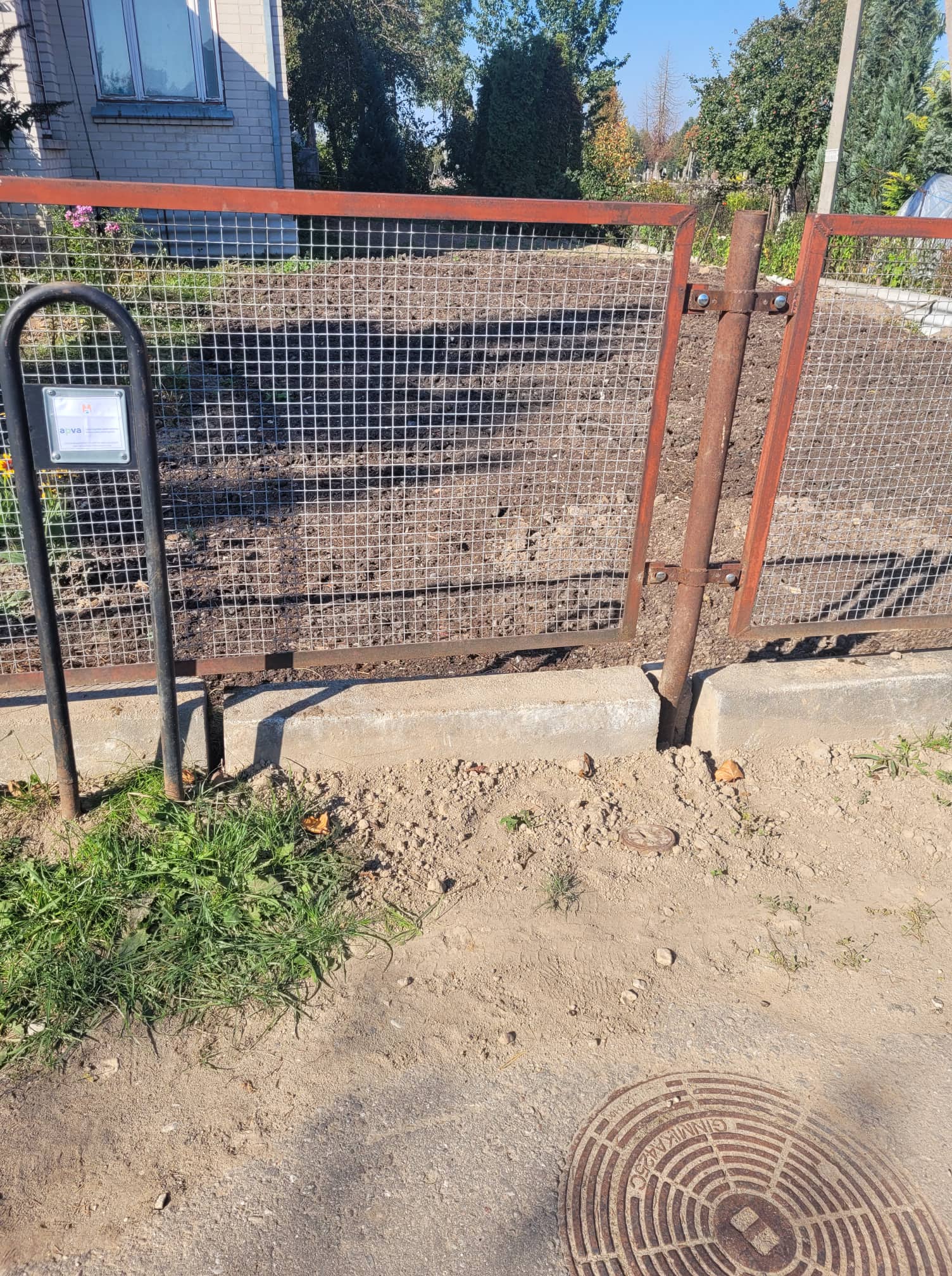 Projekto vietos nuotrauka: įrengtas nuotekų šulinys prie kiemo vartų ir finansavimo ženklas.